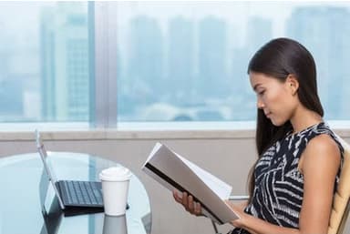 Woman in an office building reading a book at a table in front of her laptop